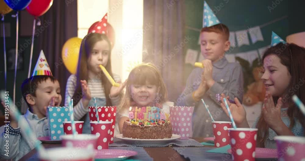 Little cute girl blowing the candles on her Happy Birthday cake and her cheerful friends standing around the table and celebrating with her at kids party. Little cute girl blowing the candles on her Happy Birthday cake and her cheerful friends standing around the table and celebrating with her at kids party.