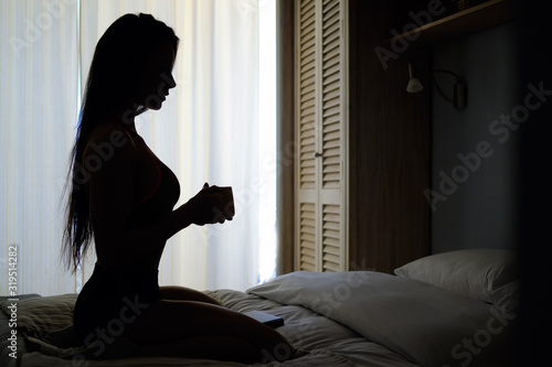 Dark silhouette of woman against window, slim young female with long hair seated on bed alone in bedroom holding morning tea cup