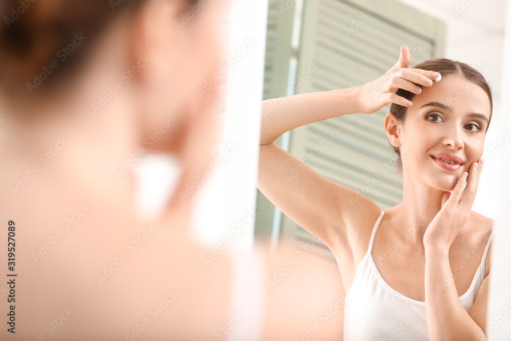Beautiful young woman applying facial cream in front of mirror
