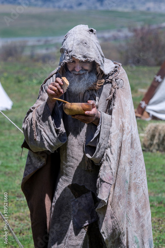 Middle-aged, bearded and ragged beggar, wandering a camp, looking for food