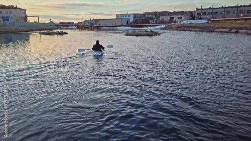 person in canoe sails along blue river leaving small waves against pictorial tundra town on banks backside view