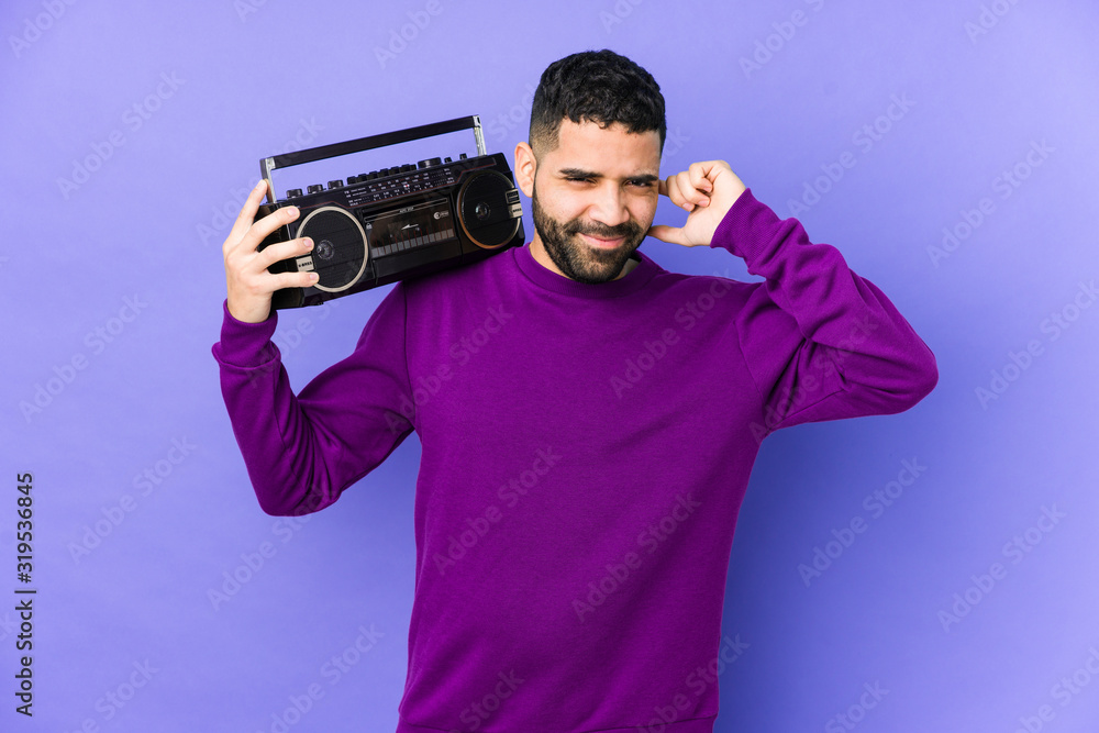 Young arabian man holding a radio cassette isolated Young arabian man listening music covering ears with hands. Young arabian man holding a radio cassette isolated Young arabian man listening music covering ears with hands.