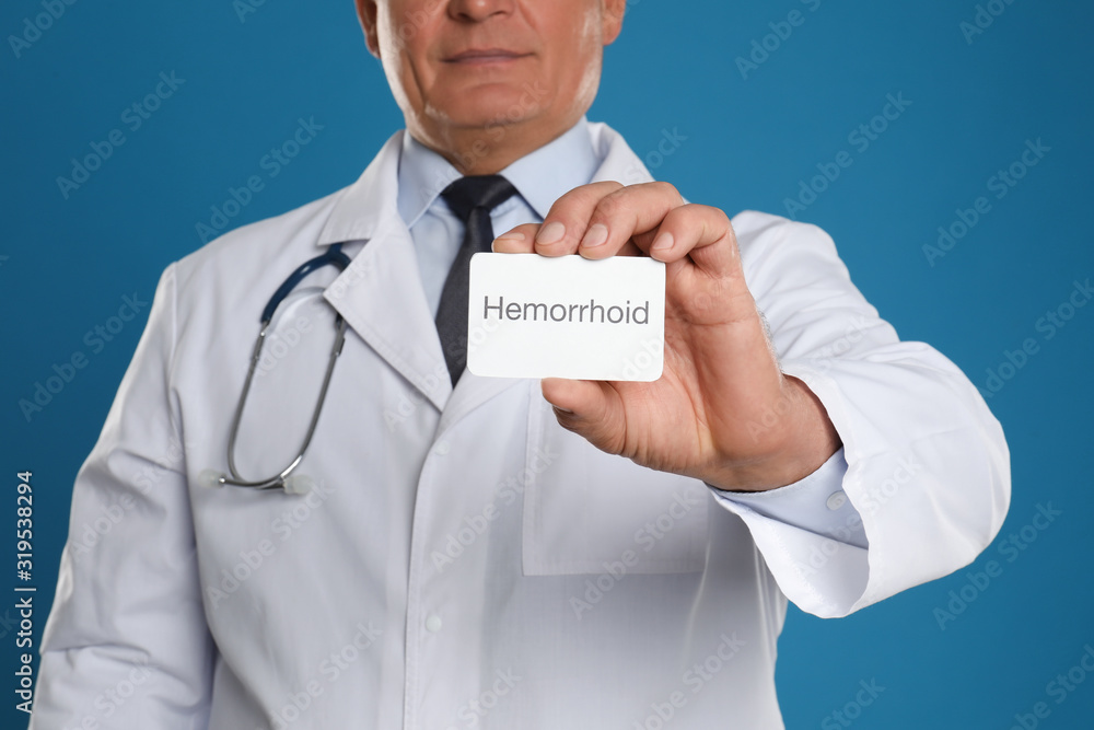 Doctor holding business card with word HEMORRHOID on blue background, closeup Doctor holding business card with word HEMORRHOID on blue background, closeup