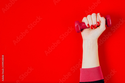A woman's hand holds a dumbbell in her fist on a bright red background. Woman power concept