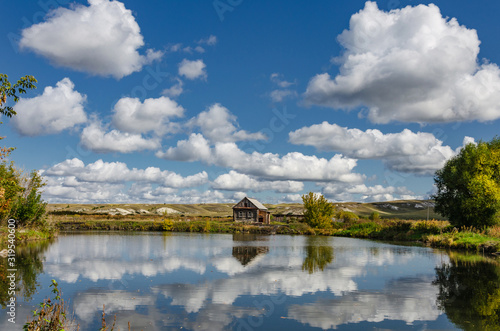 Photo of a lonely wooden house on the lake, which reflects summer clouds against the backdrop of chalk hills