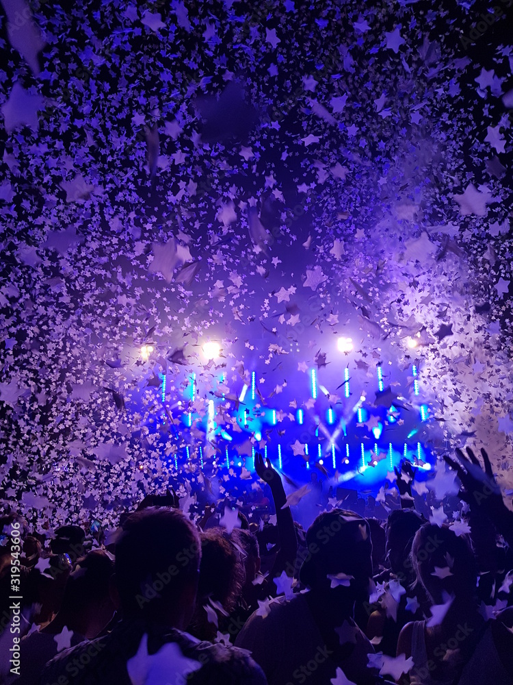 Menschen feiern mit Armen in der Luft vor einer Bühne auf einem Musikfestival mit Konfetti und bunten Scheinwerfern Menschen feiern mit Armen in der Luft vor einer Bühne auf einem Musikfestival mit Konfetti und bunten Scheinwerfern