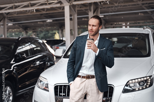 Mode of transport. Young stylish businessman in suit standing near electric car on parking drinking hot coffee looking aside confident © Viktoriia