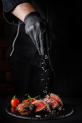 The concept of cooking meat. The chef cook salt on the cooked steak on a black background, a place under the logo for the restaurant menu. food background image, copy space text
