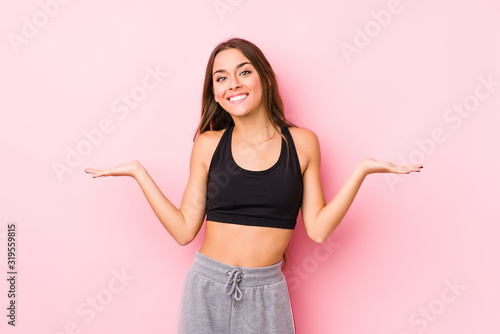 Young caucasian fitness woman posing in a pink background makes scale with arms, feels happy and confident.