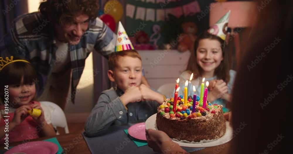 Happy and cute Caucasian kids in conuses sitting at the table at birthday party and waiting for the cake ith father while mother bringing a cake with candles. Happy and cute Caucasian kids in conuses sitting at the table at birthday party and waiting for the cake ith father while mother bringing a cake with candles.