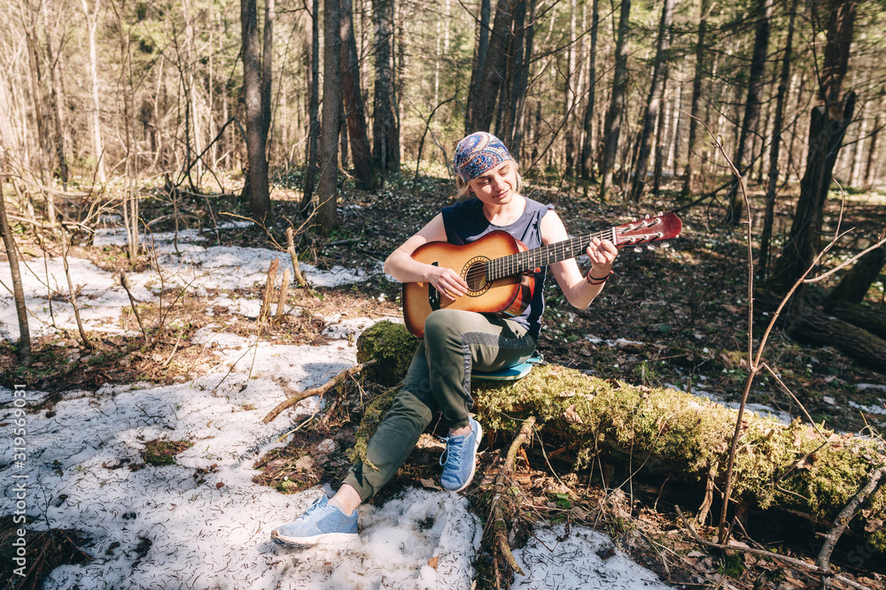 Young woman playing guitar sitting on old tree covered with moss at sunny spring forest
