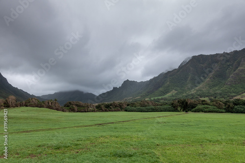 Kaaawa, Oahu, Hawaii, USA. - January 11, 2020: Kualoa valley shows deep green meadow with dirt road tearing a line and forested flanks of tall mountains on both sides under cloudy rainy sky.