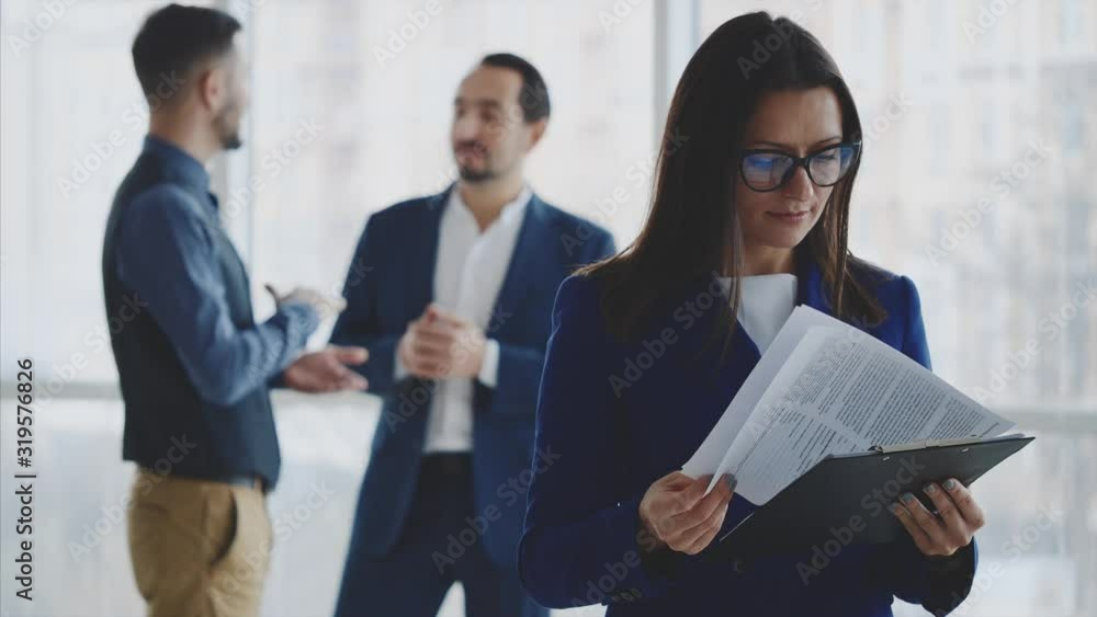 Male business workers are discussing issue on the light blurred background. Woman is turning over pages on the forefront. She is smiling, wearing glasses. Copy space. Close up. 4K. Male business workers are discussing issue on the light blurred background. Woman is turning over pages on the forefront. She is smiling, wearing glasses. Copy space. Close up. 4K.