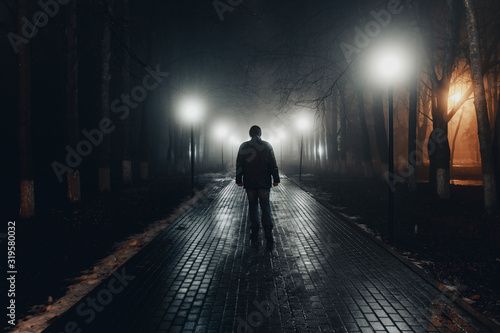Sad man alone walking along the alley in night foggy park. Back view photo