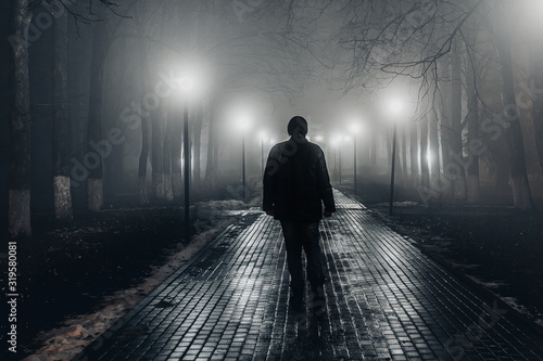Sad man alone walking along the alley in night foggy park. Back view photo
