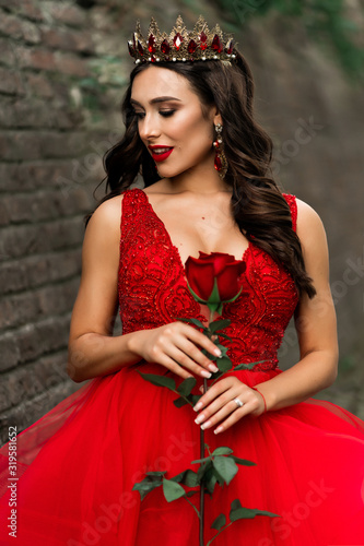 Beautiful brunette in a red dress with a crown. A brunette girl with one red rose outside on stone loft wall background. Artistic photo. Wedding photosoot photo