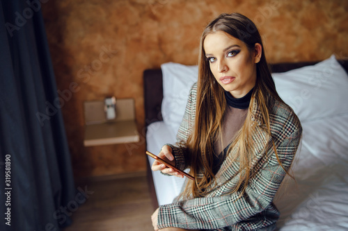 Beautiful caucasian young woman female girl tourist sitting on the bed at home or hotel apartment using mobile smart phone texting sending message app application or making a call browsing internet © Miljan Živković