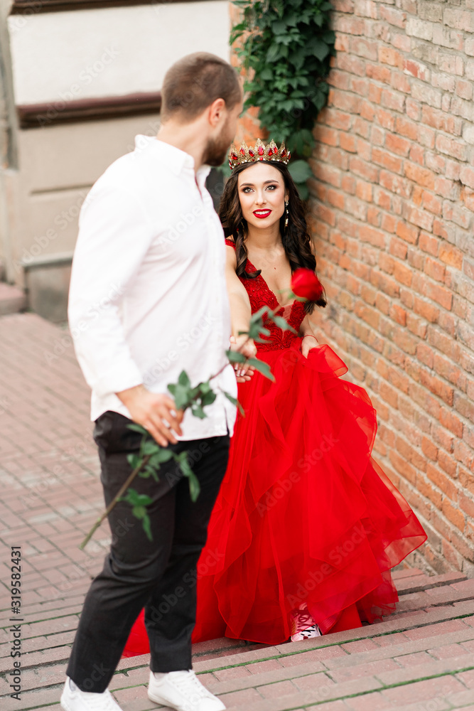 Beautiful romantic couple. Attractive young woman in red dress and crown with handsome man in white shirt with red rose walking on the street.Happy Saint Valentine's Day. Pregnant and wedding concept.