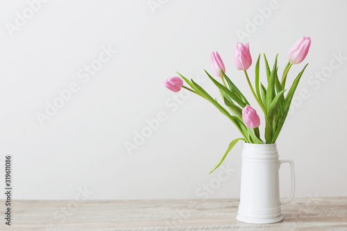 pink tulips in white ceramic jug on wooden table on background white wall