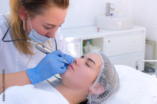 Cosmetologist making lips microblading procedure for girl using tattoo machine. Beautician is applying permanent makeup on woman's lips. Beauty industry concept. Painting lips contour.