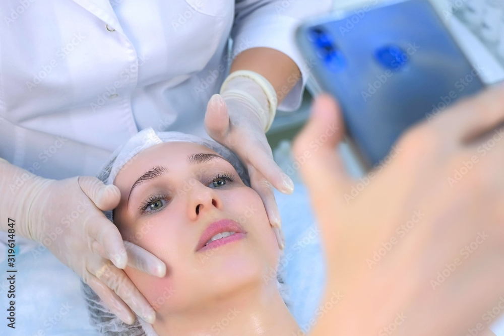 Fototapeta premium Woman blogger was making facial massage shotting video on mobile phone talking with subscribers, closeup portrait. Cosmetologist massaging mature woman face in beauty clinic. Skincare procedure.