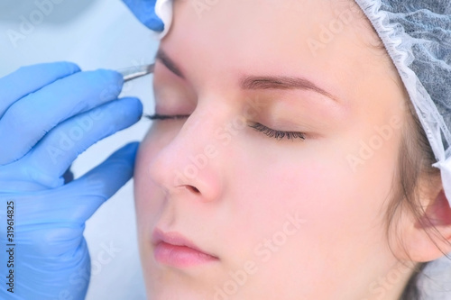 Cosmetologist plucks out hair on eyebrows with tweezers for woman, face closeup. Eyebrows tweezing procedure. Beautician is making brows correction with cosmetic tweezer. Beauty industry.