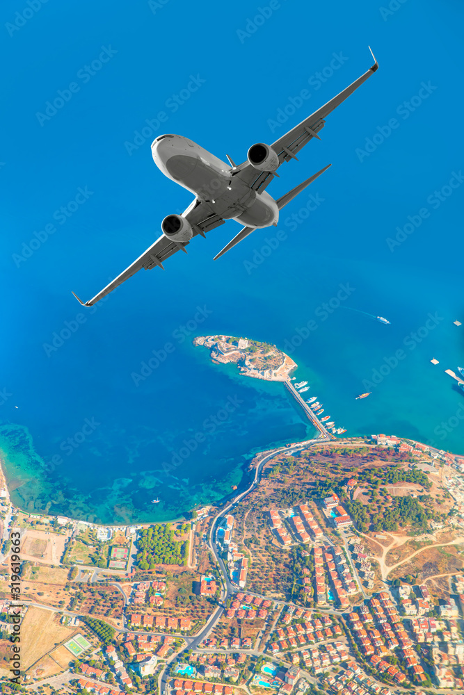 Obraz premium White passenger jet plane in the sky - Aircraft flies high over the city and sea coast of Kusadasi port and island - Kusadasi, Izmir