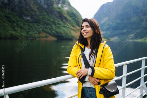 The girl tourist on a pleasure boat on the fjord enjoys the picturesque mountains and lakes of Norway. Young woman posing against the backdrop of the mountains. Travelling, lifestyle, adventure. © maxbelchenko