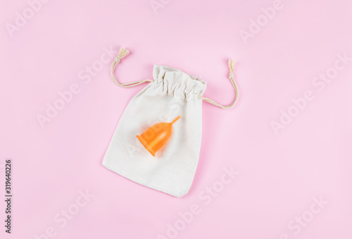 Orange menstrual cup on white cotton bag on pink background. Woman's health and environmentally friendly feminine hygiene product concept.