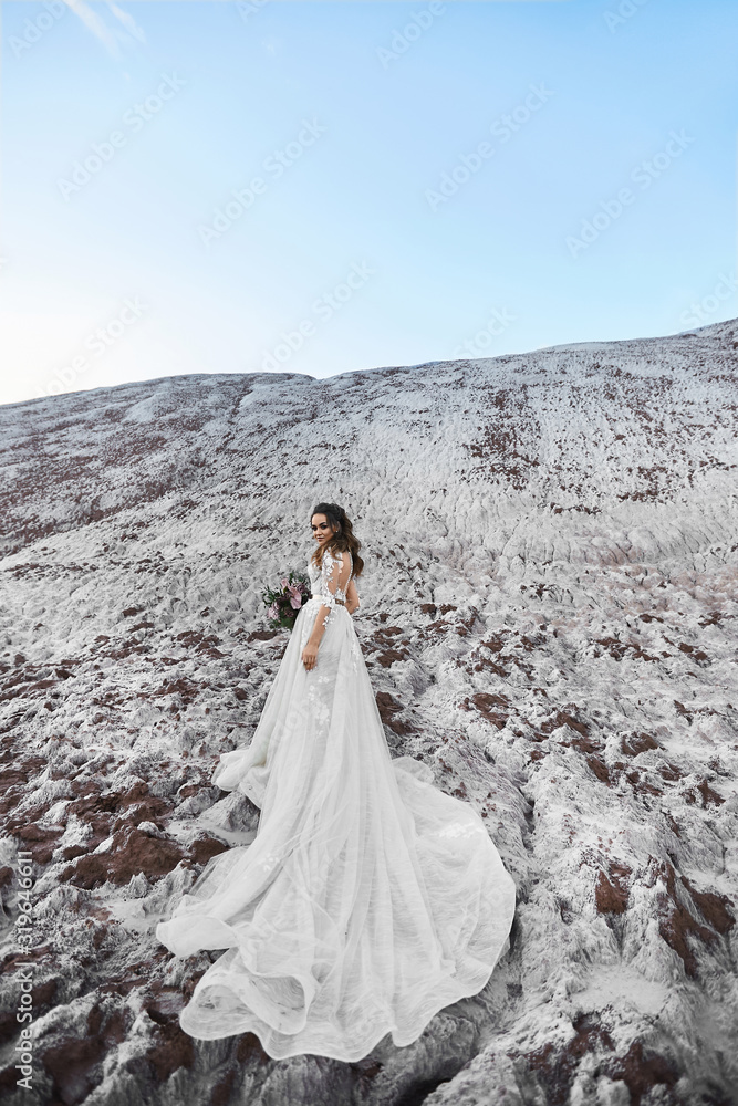 A beautiful young woman in a modern bridal dress keeping bridal bouquet and posing on a mountain with blue sky on the background on a sunny summer day. Wedding fashion concept. Copy space on the top