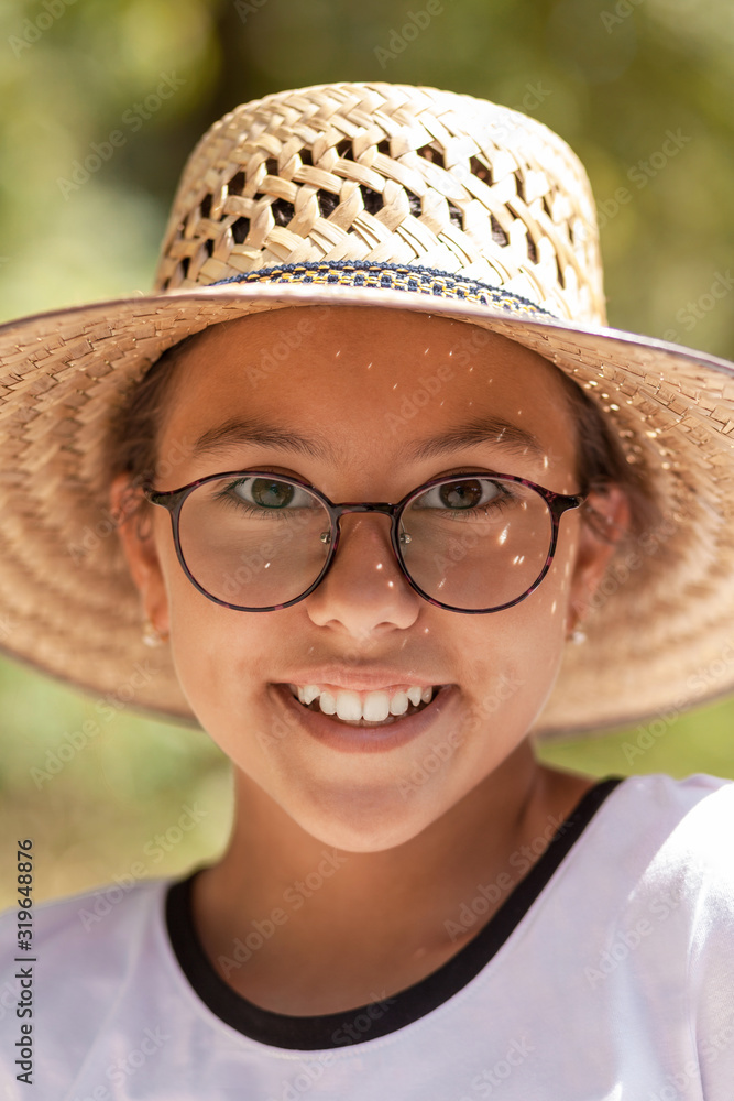 Smiling teenager girl in a straw hat and glasses. Close-up. The background is green and blurry.