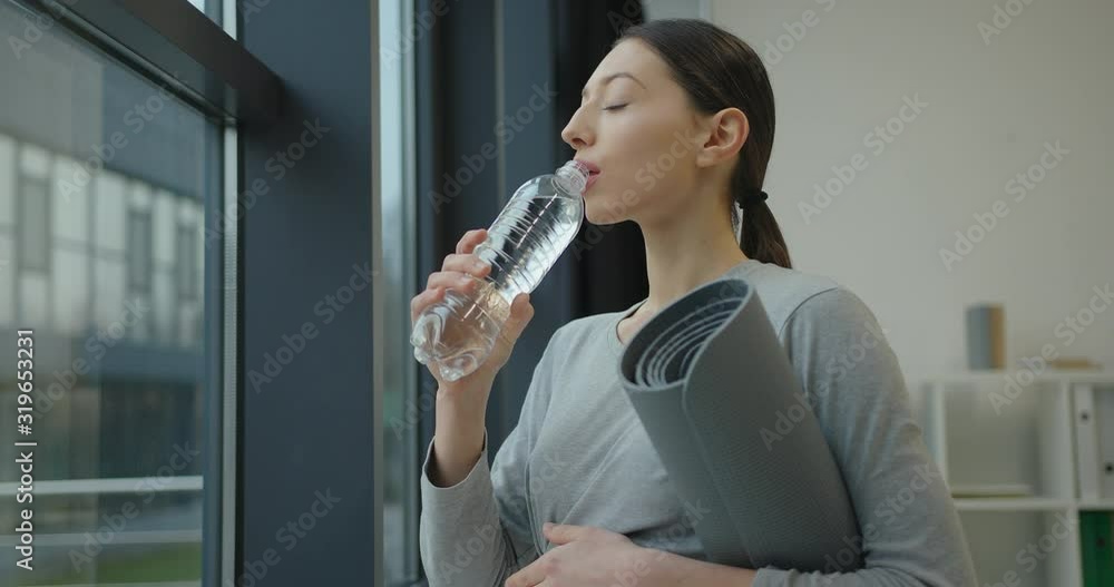 athletic young woman standing near the window, drink water and holding a gray fitness mat in her hands athletic young woman standing near the window, drink water and holding a gray fitness mat in her hands