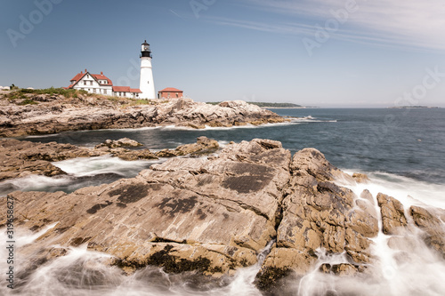 Long exposure shot of Portland Head Light, including the rough coastline and smashing waves, on a beautiful summer day.