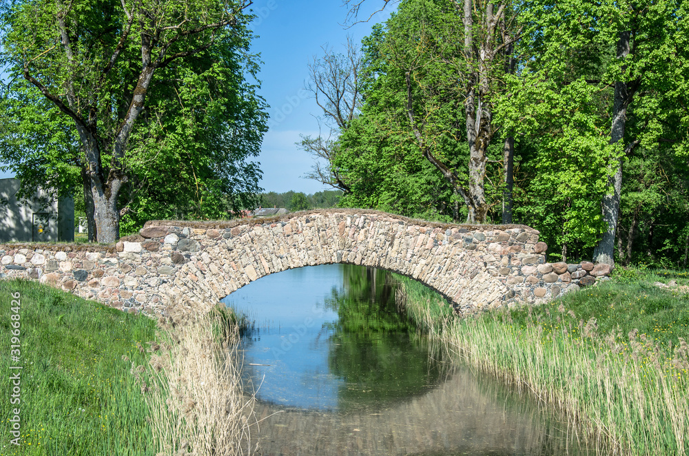 Old stone single arch bridge, Latvia. Famous ancient stone arch single track road bridge in the forest. View of small river and trees reflection in the water. Old stone single arch bridge, Latvia. Famous ancient stone arch single track road bridge in the forest. View of small river and trees reflection in the water.