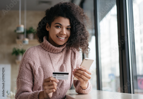 Beautiful African American woman holding credit card, making payment, looking at camera and smiling. Mobile banking concept. Young happy girl using smartphone, shopping online, booking tickets