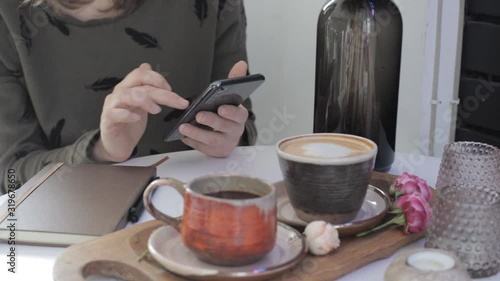 Girl holding a phone in her hands and scrolling pages while sitting at a table with coffee and a diary