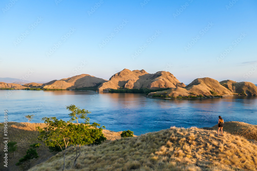 A woman standing on top of a small island, enjoying the morning sun over Komodo National Park, Flores, Indonesia. Golden hour over the islands and sea. Some boats anchored to the bay. New day 