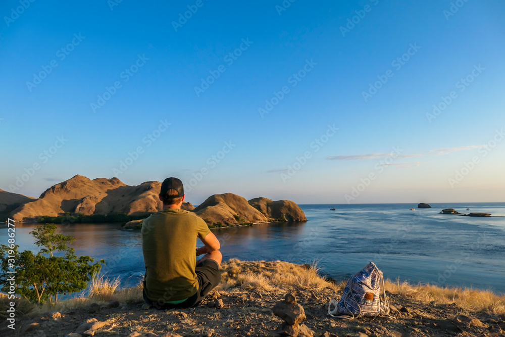A man sitting on top of a small island, enjoying the morning sun over Komodo National Park, Flores, Indonesia. Golden hour over the islands and sea. Some boats anchored to the bay. New day beginning