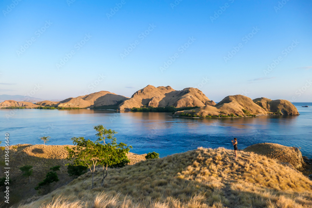 A woman standing on top of a small island, enjoying the morning sun over Komodo National Park, Flores, Indonesia. Golden hour over the islands and sea. Some boats anchored to the bay. New day 