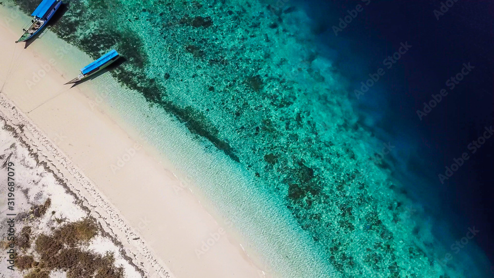 A drone shot of a small island near Maumere, Indonesia. There is a small fisherman's cottage next to the beach. Two boats anchored to the shores of the white sand beach. Calmness and solitude