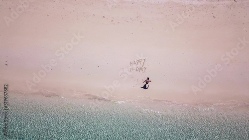 A drone shot of a man lying on a pink sand beach next to 'Happy Birthday' sign on a small island near Maumere, Indonesia. Happy and careless moments. Waves gently washing the shore. Paradise island