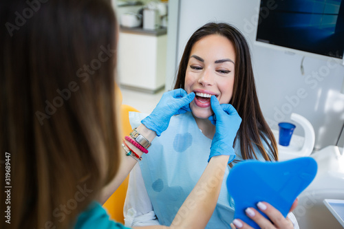 Beautiful young woman checking teeth in mirror at dentist office, showing perfect straight white teeth.