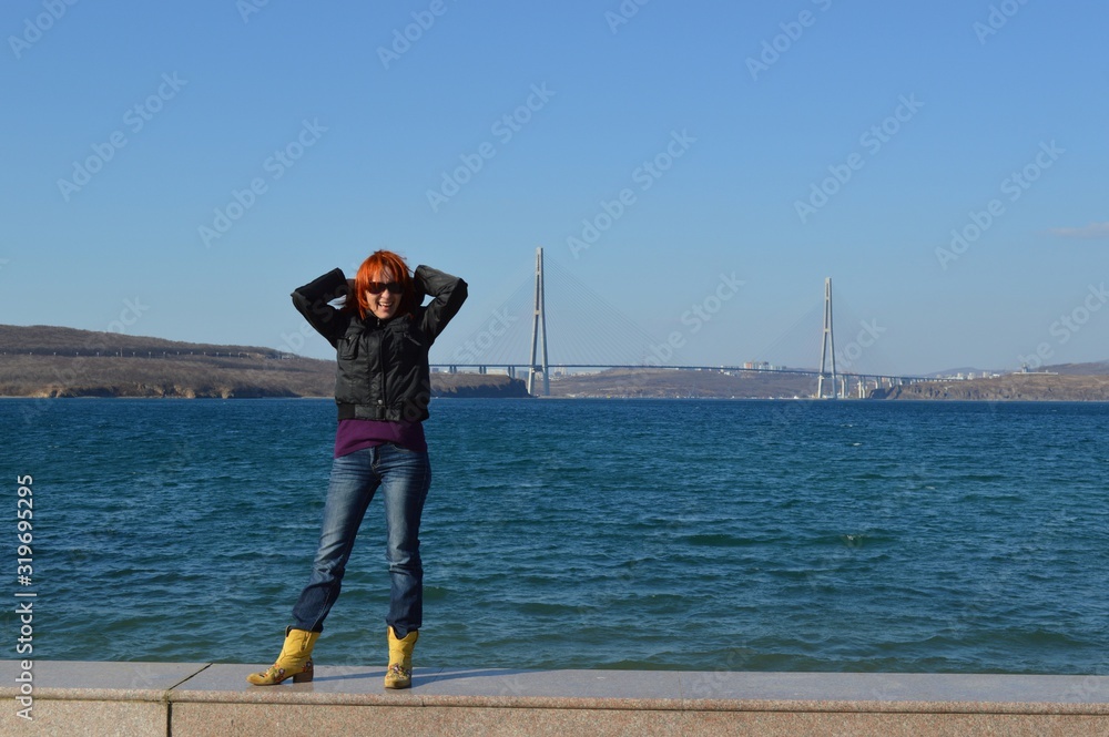 Pretty young woman with red short hair and sunglasses sincerely natural laughing and doing grimaces against the background of the sea and the Russky bridge
