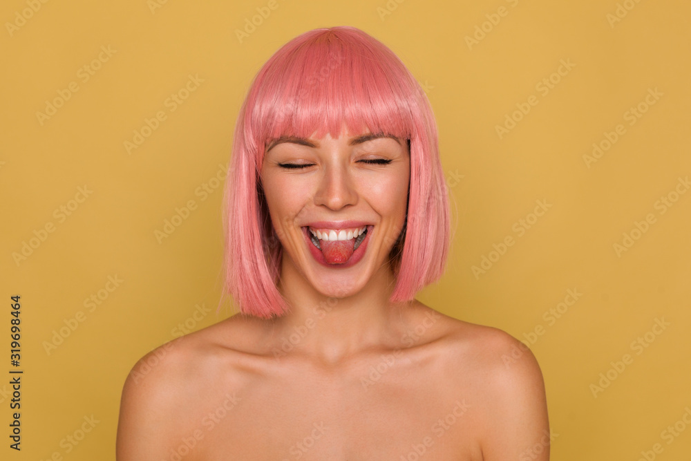 Good looking cheerful young attractive pink haired lady with natural makeup having fun while posing over mustard background, keeping eyes closed while showing happily tongue Good looking cheerful young attractive pink haired lady with natural makeup having fun while posing over mustard background, keeping eyes closed while showing happily tongue