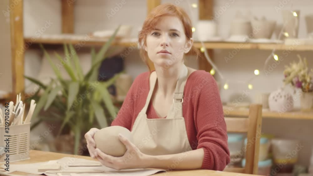 Waist-up portrait shot of young Caucasian female pottery artist with red hair, in apron sitting in ceramics studio, working on wet earthenware vessel, then looking at camera and smiling Waist-up portrait shot of young Caucasian female pottery artist with red hair, in apron sitting in ceramics studio, working on wet earthenware vessel, then looking at camera and smiling