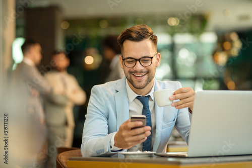 Happy young business man sitting at cafeteria with laptop and mobile. Businessman typing on smart phone while sitting in a pub restaurant. © bugarskipavle3