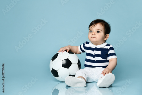 Toddler in striped t-shirt, white pants and booties. He is looking up, sitting on floor against blue background. Close up
