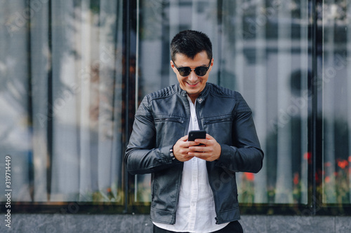 young stylish guy in glasses in black leather jacket with phone on glass background