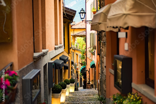 Fototapeta Naklejka Na Ścianę i Meble -  Narrow street with orange buildings and lanterns in Bellagio town at the famous Italian lake Como, Lombardy, North Italy with stone steps and Alps mountains on background.