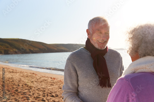 Loving Retired Couple Holding Hands Standing By Shore On Winter Beach Vacation Against Flaring Sun